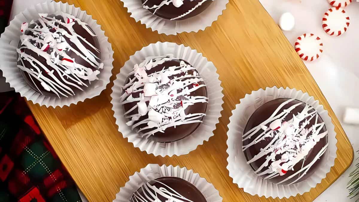 Five chocolate bombs drizzled with white chocolate and sprinkled with crushed peppermint, placed in white paper cups on a wooden board.