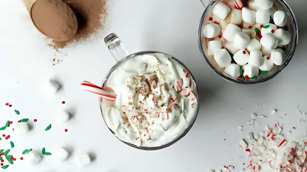 Two mugs of hot chocolate topped with whipped cream and marshmallows are shown. One has a candy cane and sprinkles, with scattered marshmallows, sprinkles, and cocoa powder around them on a white surface.