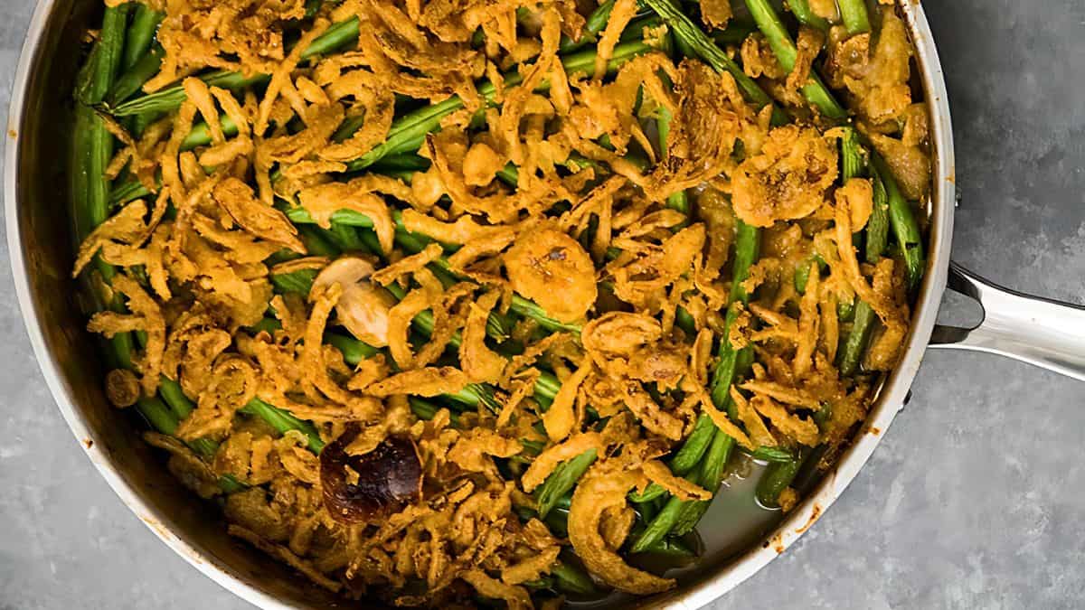 A skillet filled with a green bean casserole topped with crispy fried onions. The dish appears golden brown with visible green beans beneath the crunchy topping. The background is a gray, textured surface.