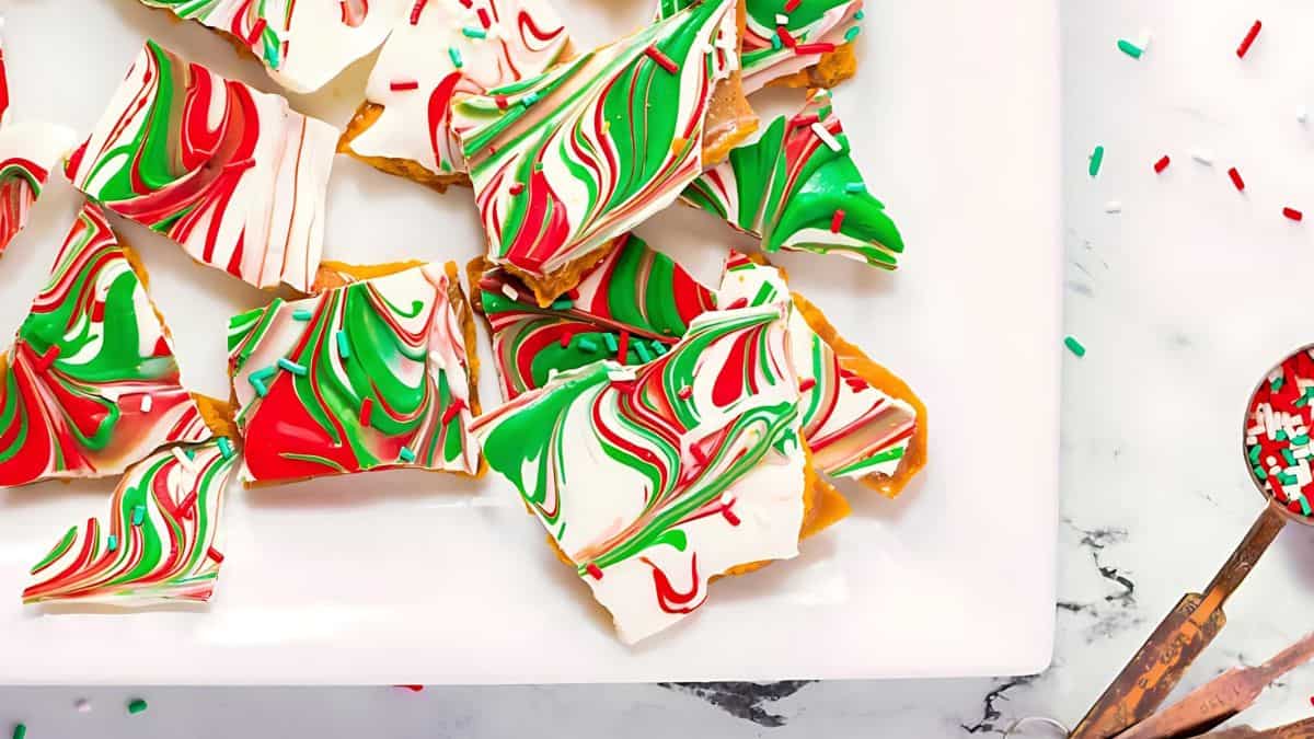 A plate of festive bark candy featuring vibrant red, green, and white swirls. The shards are artistically arranged, creating an eye-catching marbled effect. A small spoon with sprinkles is nearby on the marble surface.