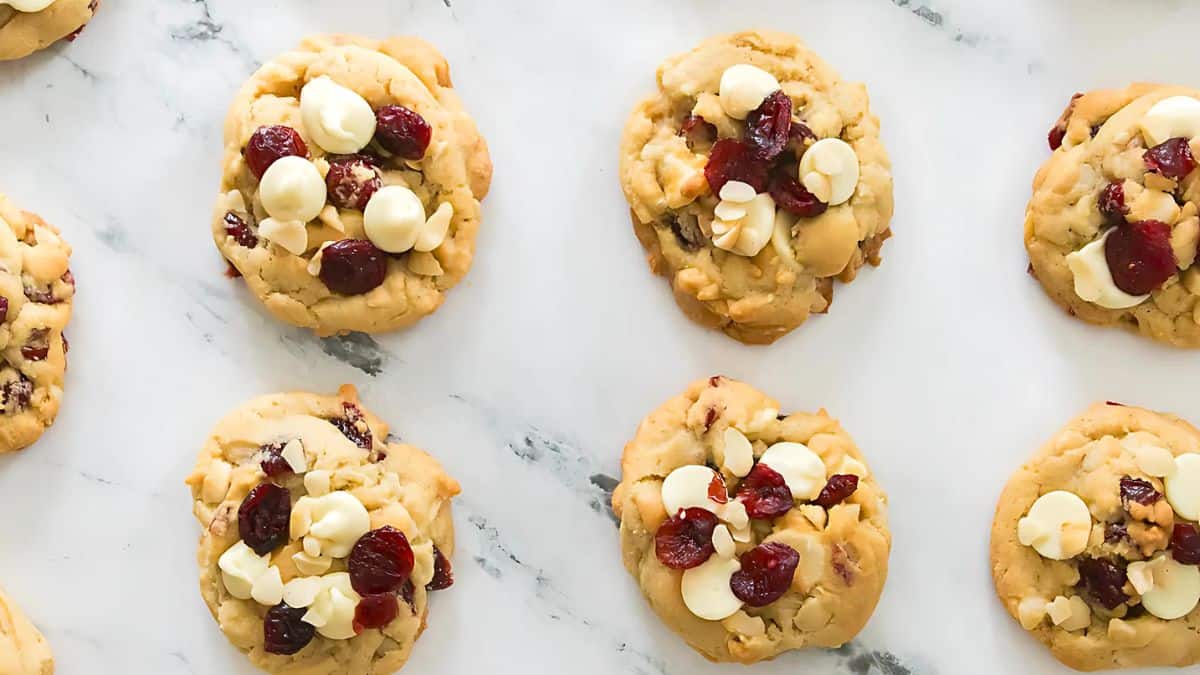 A batch of cookies with white chocolate chips and dried cranberries is displayed on a marble surface. The cookies are arranged in neat rows, showcasing their golden-brown color and textured toppings.