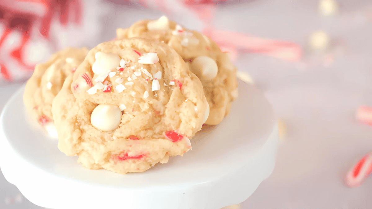 Three cookies topped with white chocolate chips and crushed peppermint candy are displayed on a white pedestal. The background is softly blurred with hints of candy canes and a light color theme.