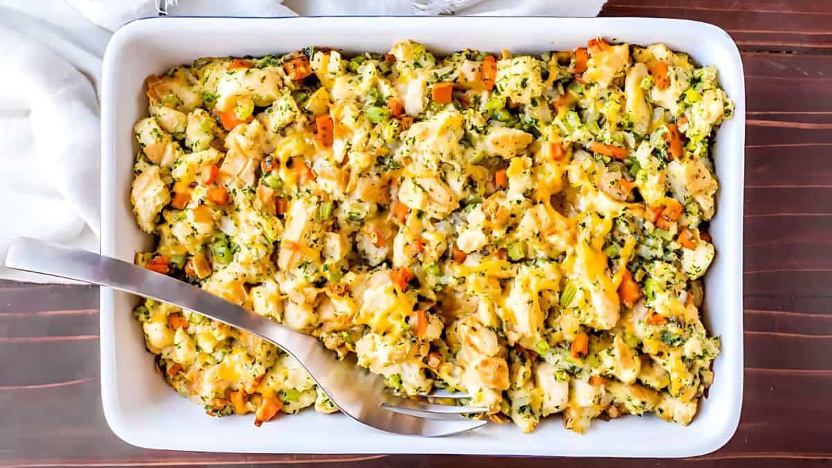 A rectangular white dish filled with a baked casserole. The dish contains pieces of chicken, broccoli, and carrots, topped with melted cheese. A serving spoon rests on the side of the dish. The dish is on a wooden surface.