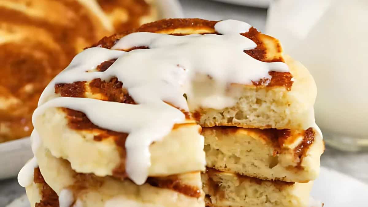 A stack of fluffy cinnamon pancakes drizzled with creamy white icing. A slice is cut out to reveal the soft interior. A glass of milk is blurred in the background.