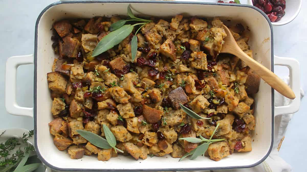 A baking dish filled with bread stuffing topped with herbs and cranberries. A wooden spoon rests on top. Fresh sage leaves are garnished on the dish. A small bowl of cranberries is visible in the corner.