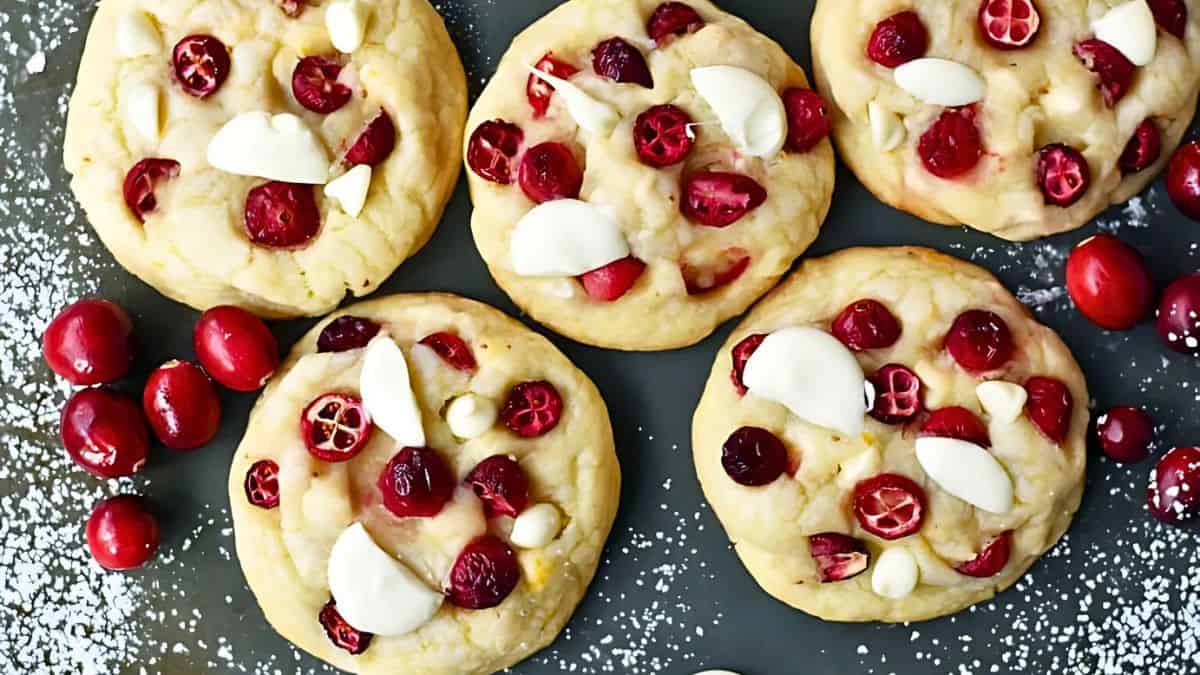 Five cookies topped with cranberries and white chocolate chunks are arranged on a dark surface with scattered cranberries and powdered sugar.