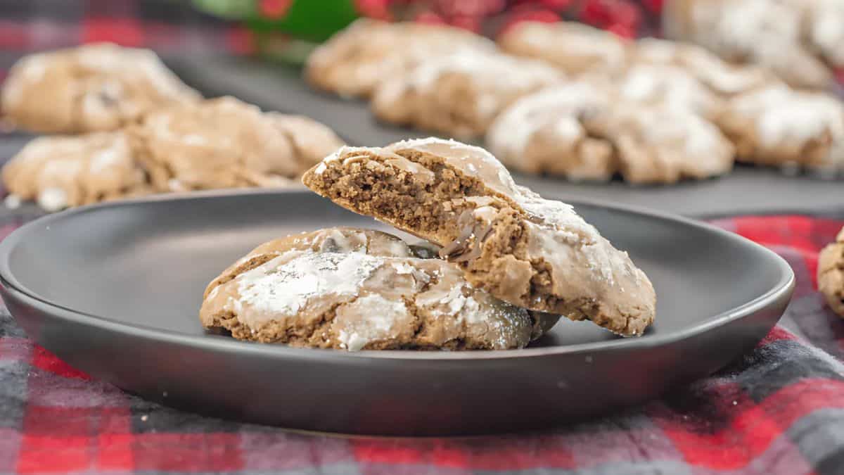 A close-up of chocolate cookies dusted with powdered sugar on a dark plate. The cookies have a soft texture, some broken open to reveal a chewy center. The background shows more cookies on a dark surface. The setup rests on a red and black plaid cloth.