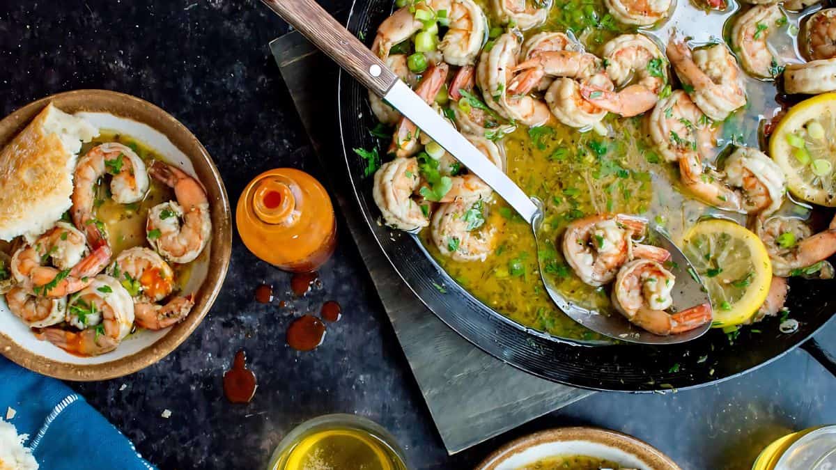 A skillet filled with shrimp in a garlic lemon sauce, garnished with herbs. A serving spoon rests in the skillet. Nearby, a plate with shrimp, a slice of bread, a bottle of hot sauce, and two glasses with drinks are visible on a dark textured surface.