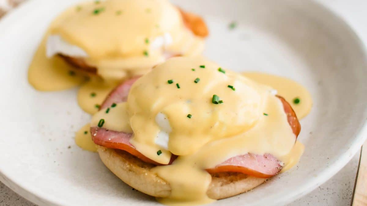 A plate with two eggs Benedict, featuring poached eggs, slices of ham, and creamy hollandaise sauce on toasted English muffins. Garnished with chopped chives.
