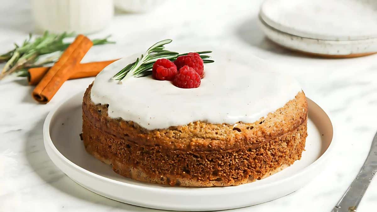 A frosted cake sits on a white plate, garnished with fresh raspberries and a sprig of rosemary. Cinnamon sticks are placed in the background on a marble surface.