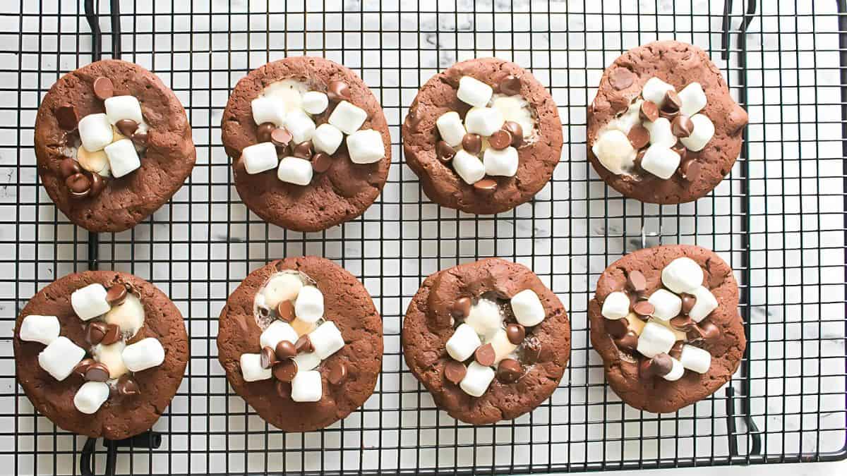 Eight chocolate cookies topped with marshmallows and chocolate chips are arranged neatly on a cooling rack. The cookies are set against a marble surface.