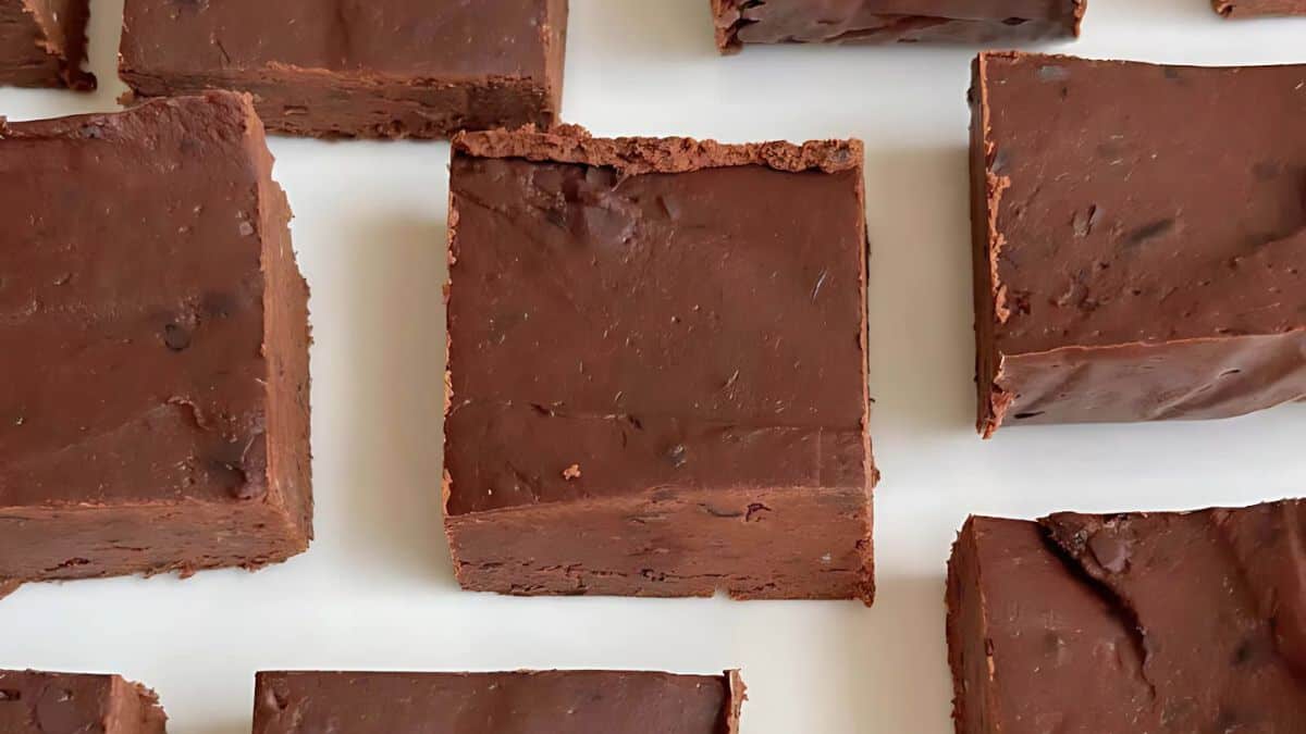 A close-up of several square pieces of rich, fudge chocolate placed on a white surface. The texture appears smooth with slightly uneven edges, and the chocolate has a glossy finish.