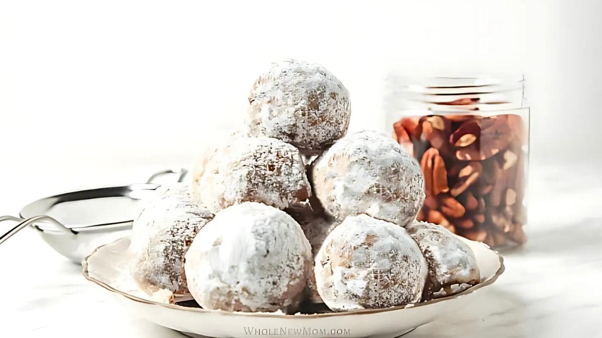 A plate of round, powdered sugar-covered cookies stacked in a pyramid shape. Nearby, there's a jar filled with almonds and a metal scoop, all set against a light background.