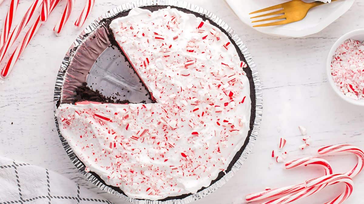 A pie with a chocolate crust and white whipped topping, sprinkled with crushed candy canes. A slice is missing. Candy canes and a plate with forks are arranged around the pie on a light wooden surface.