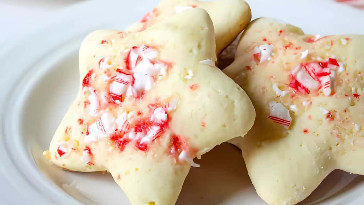 Star-shaped cookies with a creamy icing are sprinkled with crushed peppermint candies, resting on a white plate.