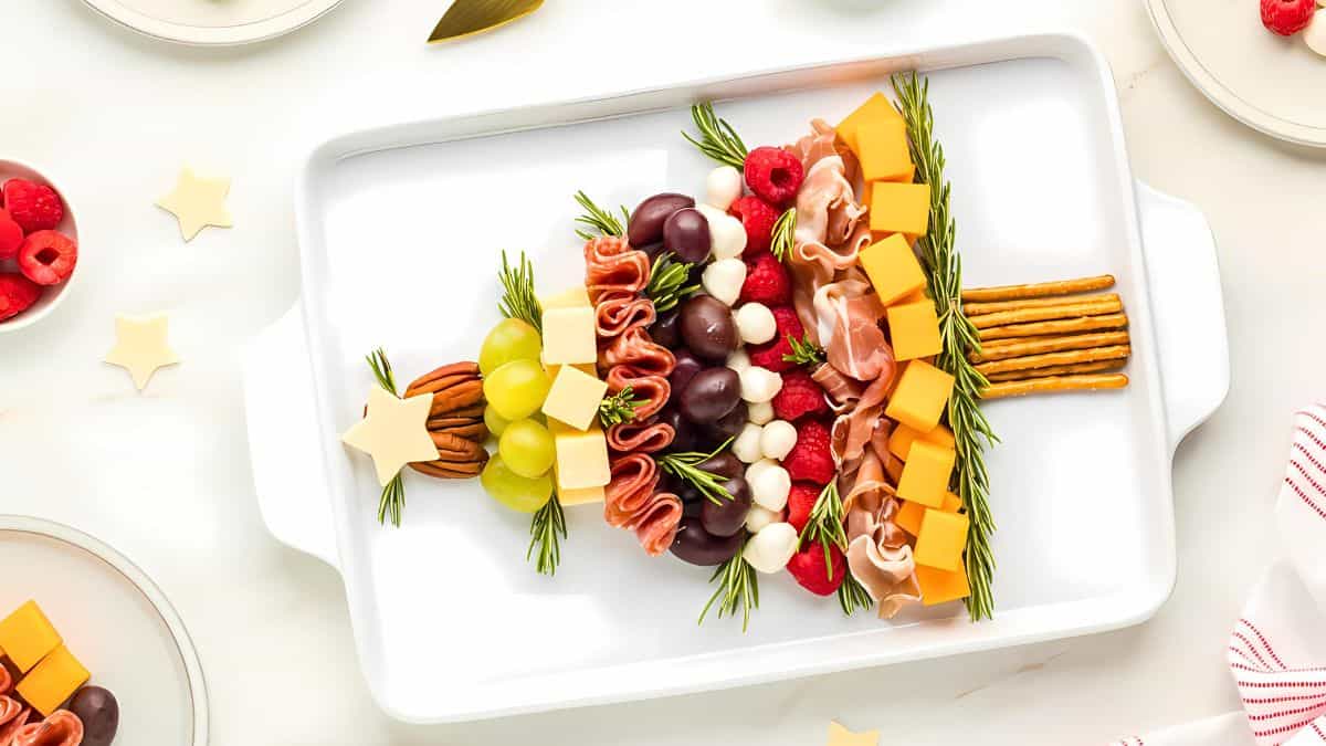 Festive appetizer shaped like a Christmas tree on a white platter, with grapes, cheese cubes, raspberries, meats, mozzarella balls, pretzels, pecans, and rosemary.