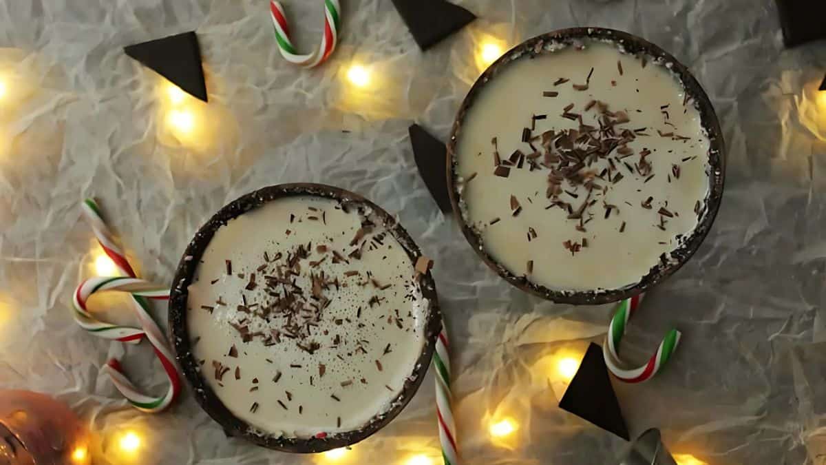 Two cups of creamy beverage topped with chocolate shavings are surrounded by festive candy canes, warm string lights, and dark chocolate triangles on a crumpled white cloth.