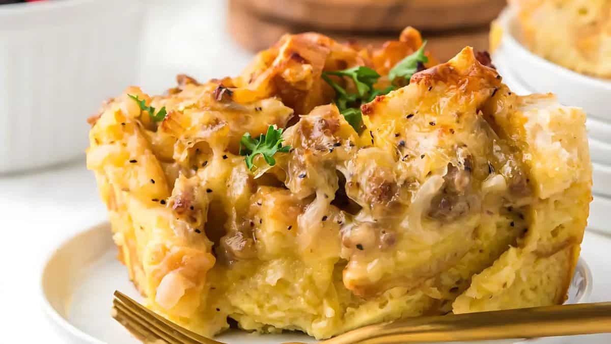A close-up of a savory breakfast casserole featuring golden, crispy edges and a creamy interior. The top is garnished with fresh parsley, and a fork rests on the white plate beside it.