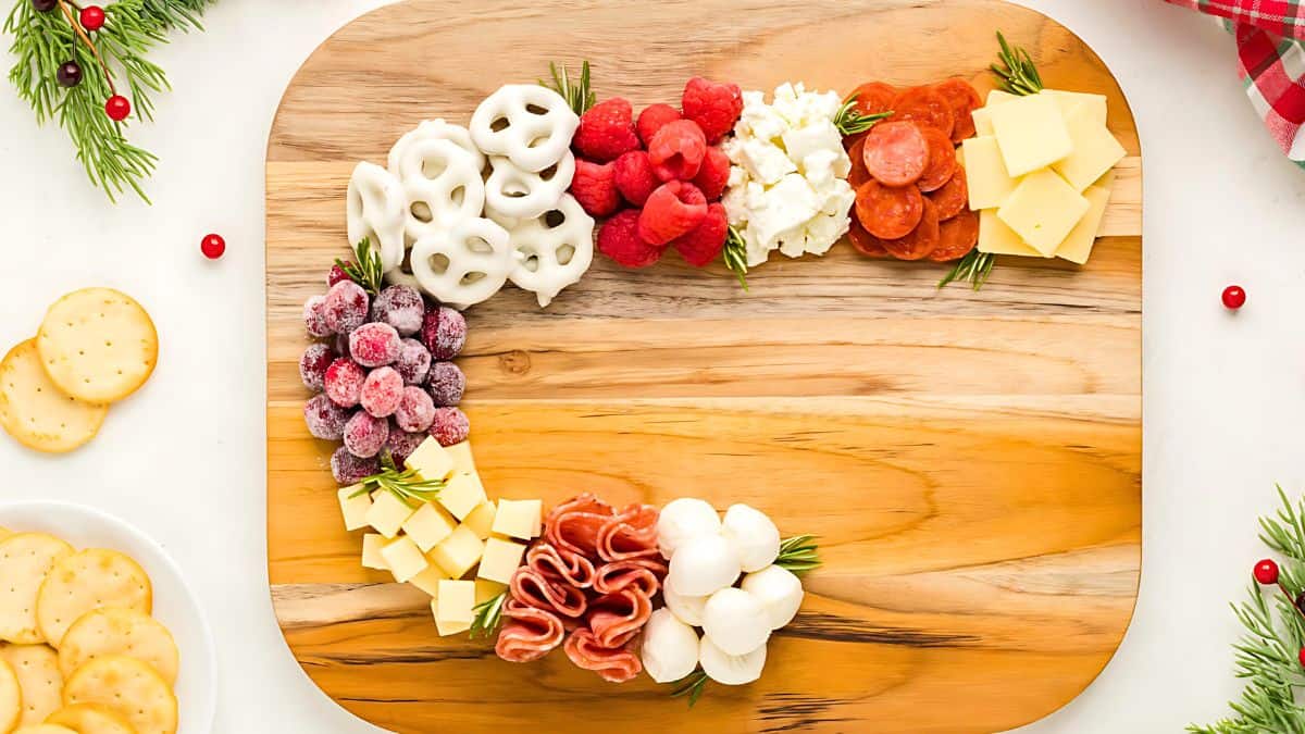 A Christmas wreath charcuterie board with white chocolate pretzels, raspberries, cheese cubes, frosted cranberries, rosemary, pepperoni, mozzarella balls, and slices of salami arranged in a circular pattern on a wooden board. Crackers are nearby.