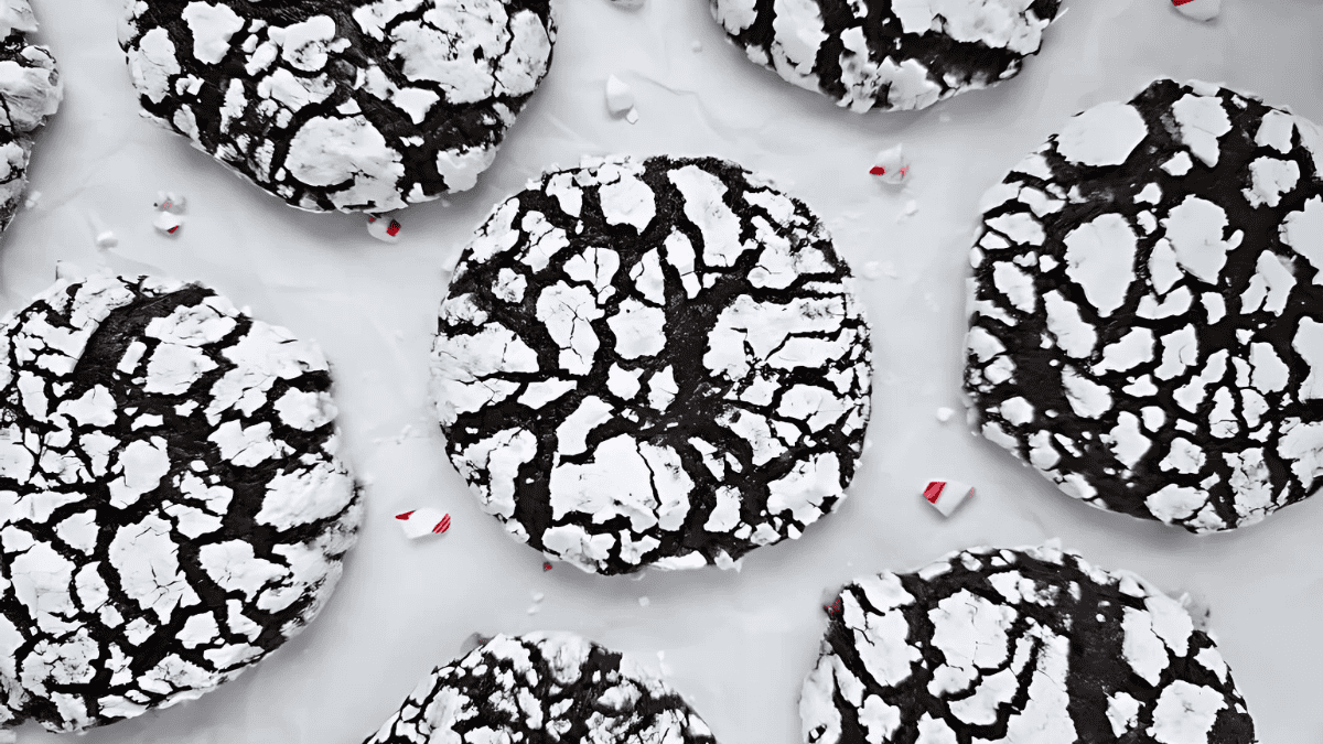Close-up of chocolate crinkle cookies with powdered sugar on top, creating a cracked pattern. The cookies are arranged on a light-colored surface, with a few small candy cane pieces scattered around.