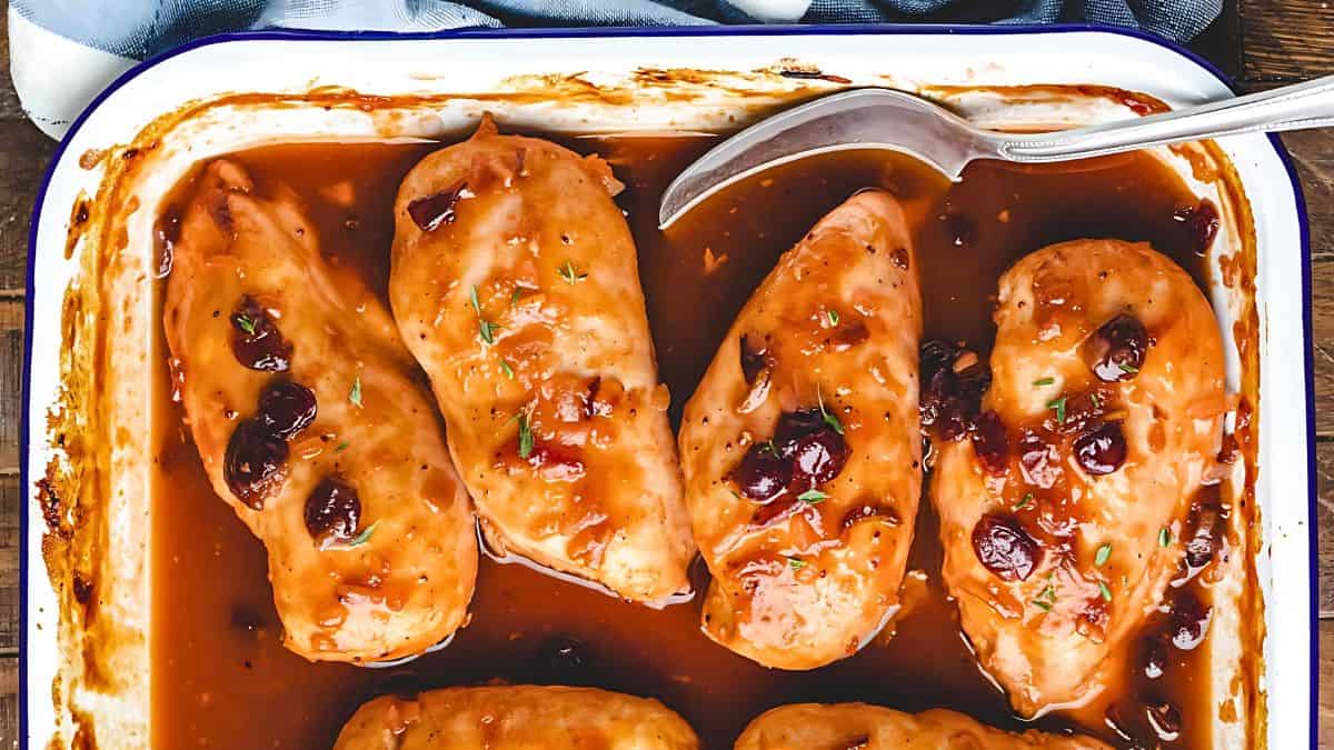 Baked chicken breasts in a rectangular dish are covered with a glossy cranberry glaze, garnished with herbs. A silver serving spoon rests on the side of the dish, which is set on a wooden table.