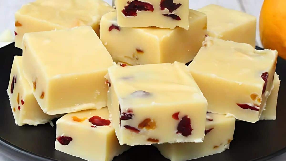 Cubes of white chocolate fudge with bits of dried cranberries and nuts are stacked on a black plate. The creamy texture and deep red fruit pieces create an inviting and festive appearance.