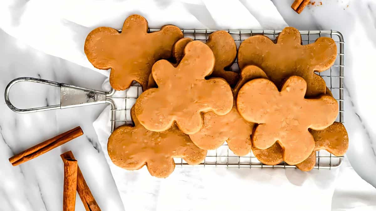 A wire rack with six gingerbread cookies shaped like gingerbread people, glazed with icing. The rack is on a white surface, and there are cinnamon sticks nearby.