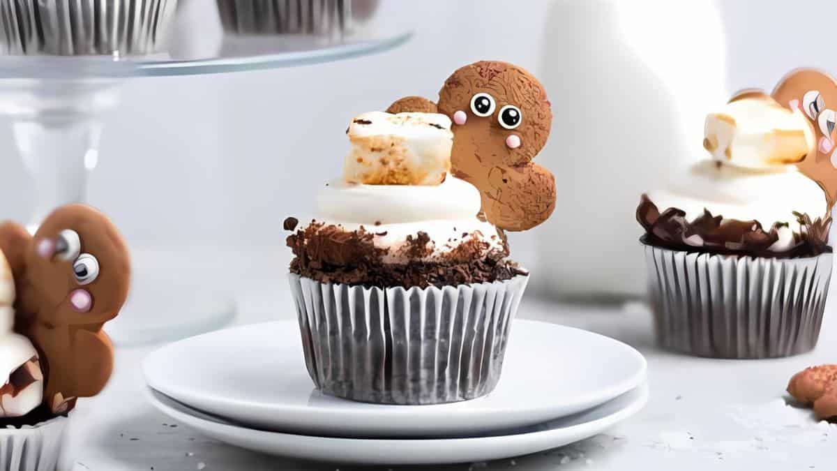 Cupcakes topped with whipped cream and toasted marshmallows are decorated with gingerbread cookies featuring cute faces. They are placed on a white plate with additional cupcakes and a bottle of milk in the background.