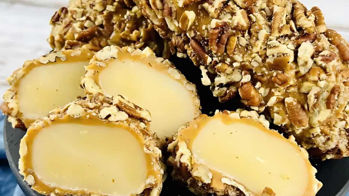 Close-up of several caramel nut log candies, coated in chopped pecans and sliced to reveal a creamy, light yellow filling. The candies are arranged on a dark plate, showcasing their rich texture and inviting appearance.