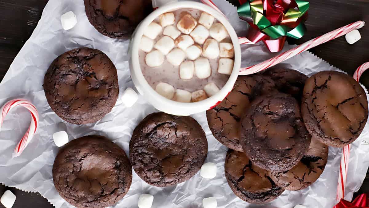 A cozy setup featuring a mug of hot chocolate topped with marshmallows, surrounded by chocolate cookies, mini marshmallows, candy canes, and a festive bow on a parchment paper background.