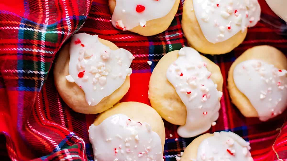 A close-up of cookies with white icing and red sprinkle decorations, placed on a red and green plaid cloth. The cookies are arranged in a festive manner, suggesting a holiday theme.
