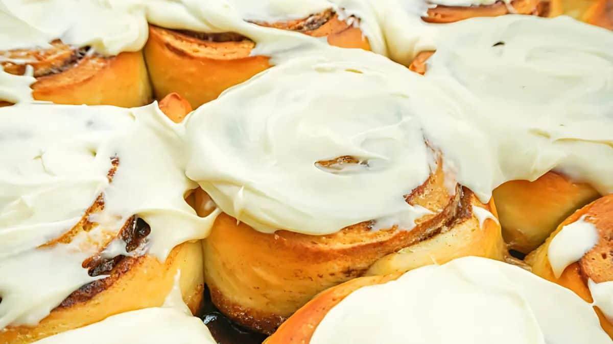 Close-up of several freshly baked cinnamon rolls topped with creamy white frosting. The rolls are arranged closely together, highlighting their swirled pastry layers and rich texture.
