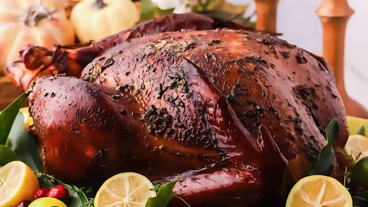 A whole roasted turkey garnished with herbs is surrounded by lemon slices and greens. Small pumpkins and candles are in the background, creating a festive atmosphere.