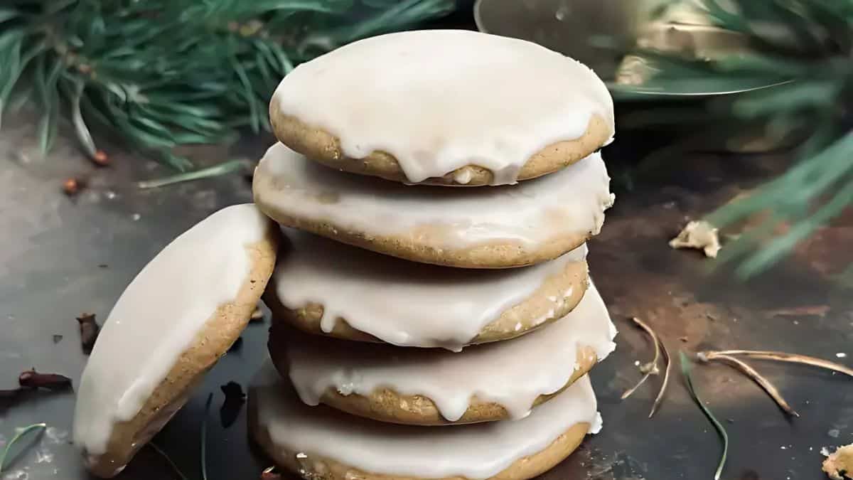 A stack of iced cookies sits on a dark surface, surrounded by pine branches and a few cloves. The cookies are lightly frosted, with some icing dripping over the edges. The setting evokes a cozy, festive atmosphere.