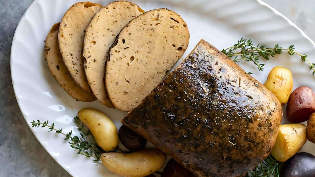 Sliced vegan roast on a white platter, garnished with herbs and surrounded by small roasted potatoes. The roast has a seasoned, herb-coated crust.