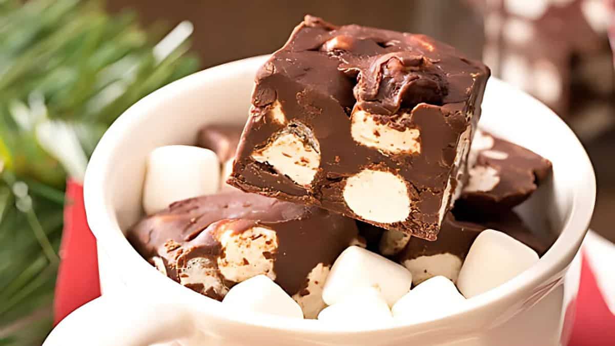 A white mug filled with cubes of chocolate fudge. Some fudge pieces feature visible marshmallows mixed in. The mug is surrounded by small marshmallows and sits on a red cloth with a hint of greenery in the background.