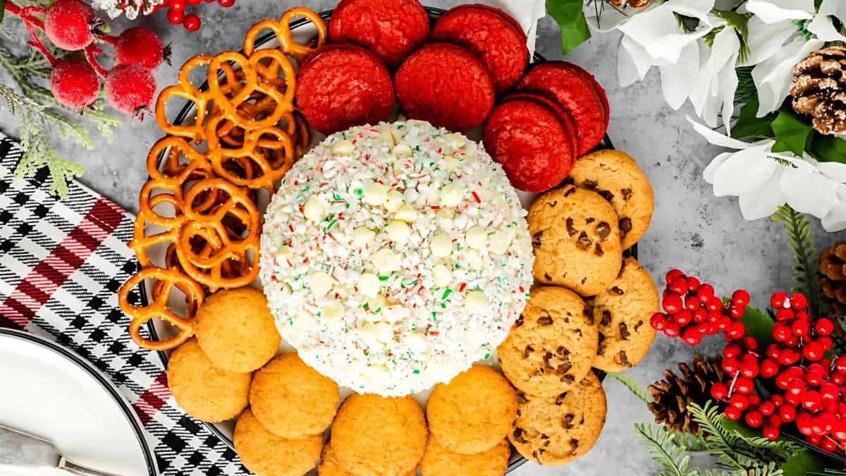 A festive arrangement of cookies and pretzels surrounds a large, sprinkle-covered cheese ball. The display is accented with winter greenery, pine cones, and red berries, set on a decorative cloth.