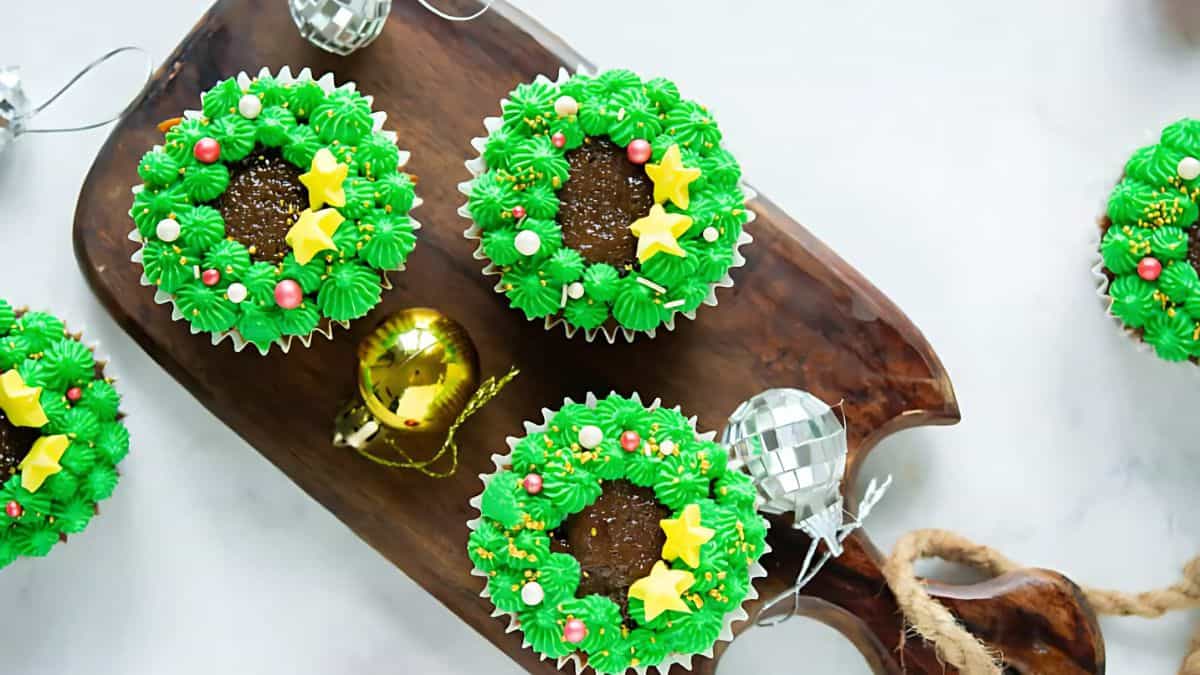 Cupcakes decorated with green frosting to resemble Christmas wreaths, topped with yellow star and red ball sprinkles, are arranged on a wooden board. Shiny silver and gold baubles accessorize the festive setting.