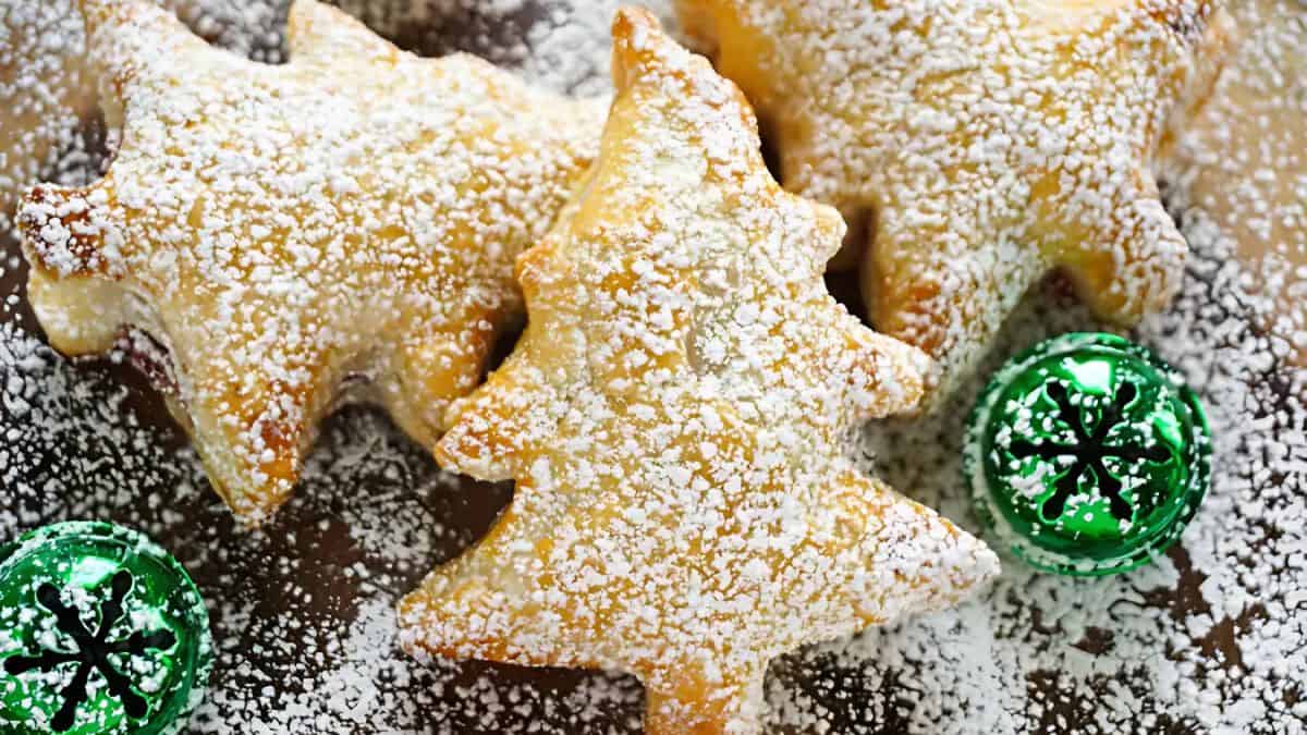 Three Christmas tree-shaped pastries are dusted with powdered sugar. Two green, metallic-wrapped chocolates with snowflake patterns are placed nearby on a wooden surface.