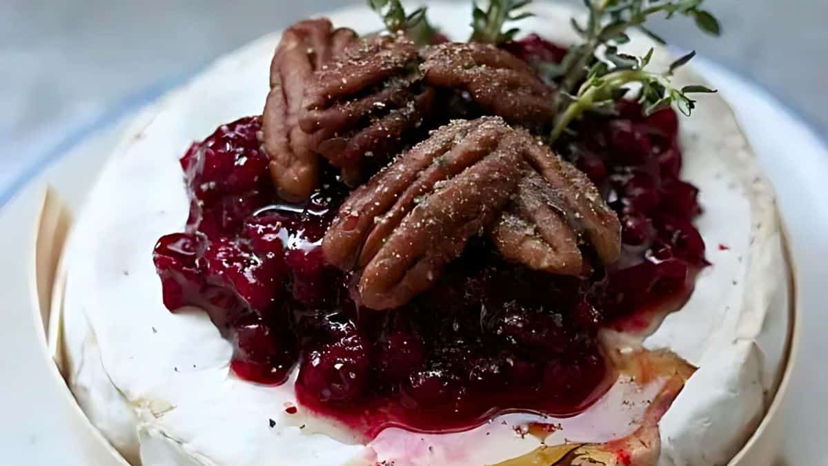 A round of baked brie topped with a layer of berry sauce, pecans, and garnished with sprigs of fresh thyme. The creamy cheese is slightly melted, with a light golden crust around the edges.
