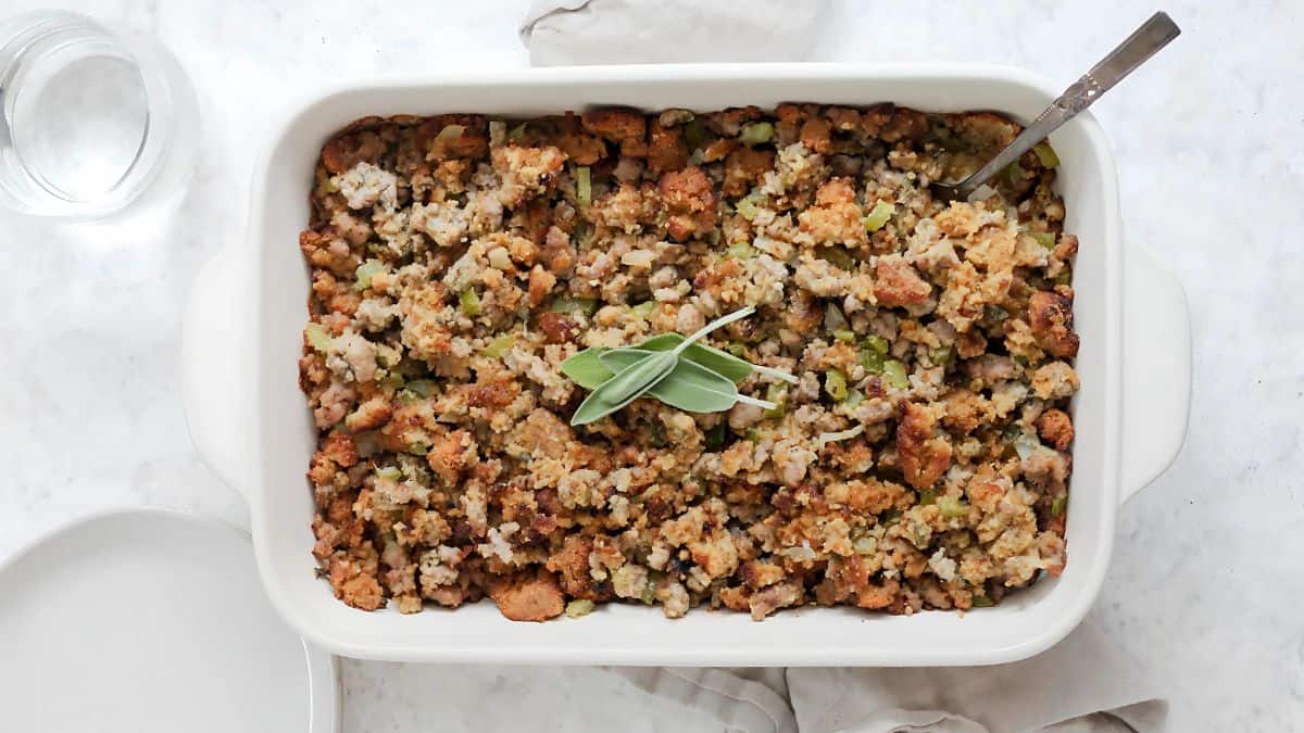 A white casserole dish filled with golden-brown stuffing, garnished with a sprig of fresh sage. A spoon rests in the dish, suggesting it's ready to serve.