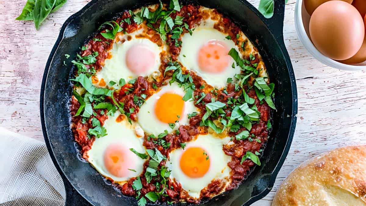 A cast iron pan filled with shakshuka, featuring five poached eggs nestled in a spiced tomato sauce. The dish is garnished with fresh herbs. Next to the pan are a bowl of eggs and a piece of rustic bread.