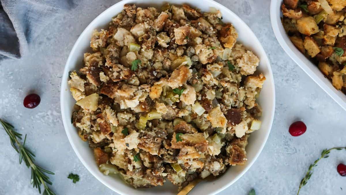 A bowl of bread stuffing with herbs, celery, and onion on a grey surface. Sprigs of rosemary and scattered cranberries surround the bowl. A second dish of stuffing is partially visible in the background.