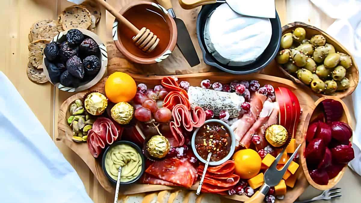 A wooden platter filled with an assortment of food including sliced meats, cheese, olives, figs, grapes, honey, mustard, Ferrero Rocher chocolates, pistachios, sliced bread, and orange slices, with a cheese knife and fork set nearby.
