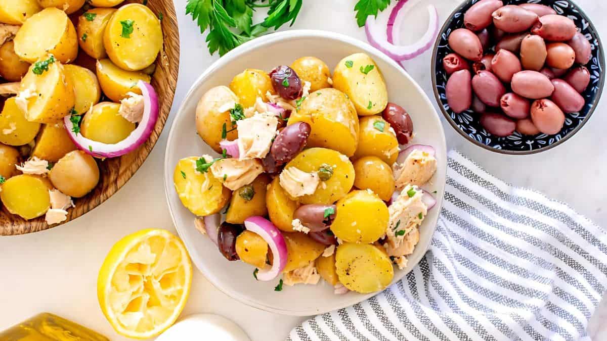 A plate of potato and olive salad with sliced red onions and herbs is presented on a white surface. Next to it, there is a bowl of olives, a leafy garnish, a striped napkin, and a halved lemon.