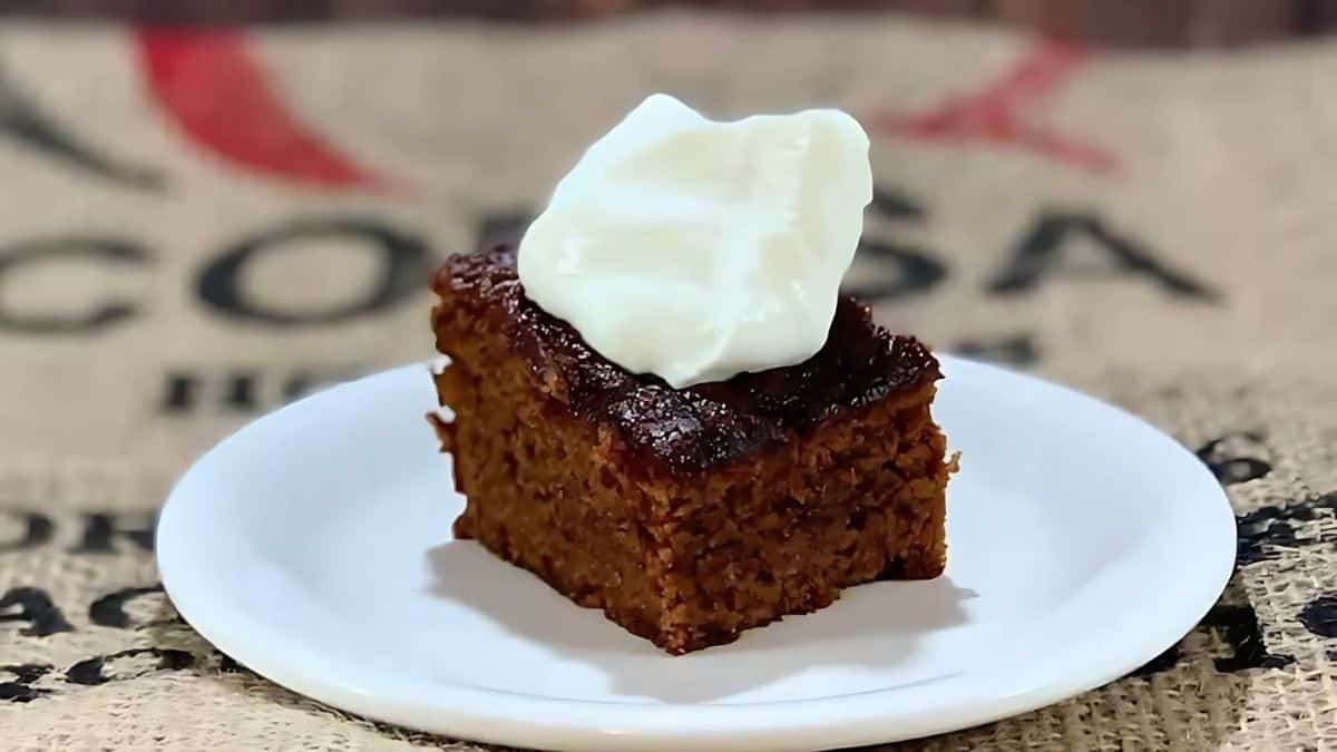 A slice of moist brown cake topped with a dollop of white cream sits on a white plate. The background features textured fabric with black and red text.