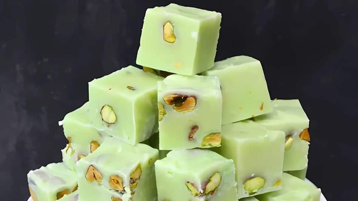 A pyramid of green pistachio fudge pieces is stacked on a dark background. Each piece is square with visible pistachio nuts embedded in the creamy, light green surface.
