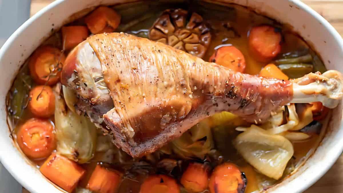 A roasted turkey leg rests atop a bed of roasted carrots, onions, and garlic in an oval dish. The vegetables are caramelized, adding a rich, warm color to the dish.