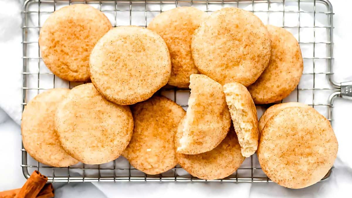 A batch of round snickerdoodle cookies is arranged on a wire cooling rack. The cookies are golden brown and dusted with cinnamon sugar. One cookie is broken in half, showing a soft interior. The scene is set against a light background.
