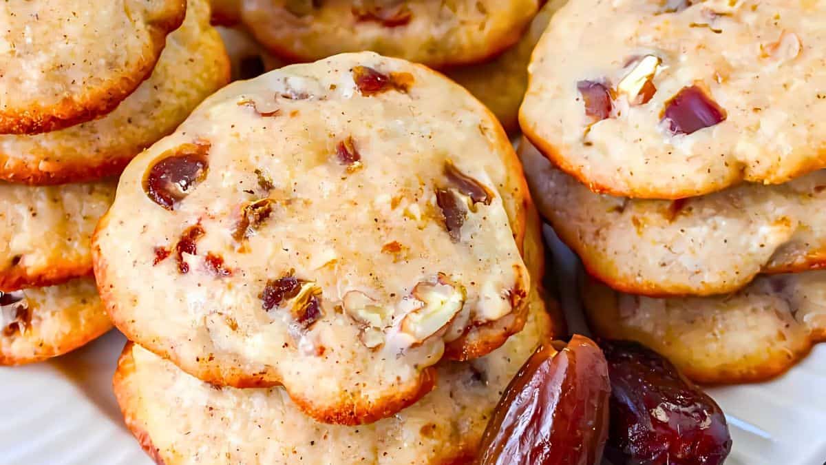 A close-up of stacked date nut cookies with visible chunks of dates and nuts. The cookies are golden brown and arranged on a white plate, with a few whole dates in the foreground.